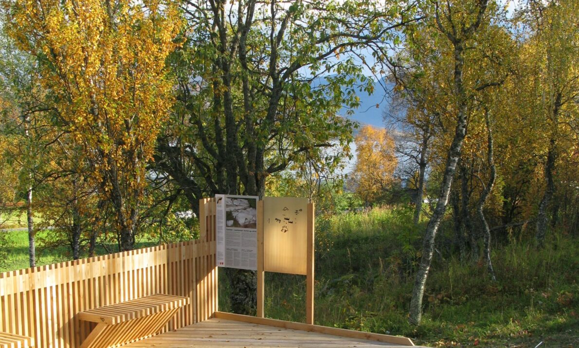 Utsiktsplattform i tre omgitt av høstfargede trær, med informasjonsskilt og fjell og vann synlig i bakgrunnen.