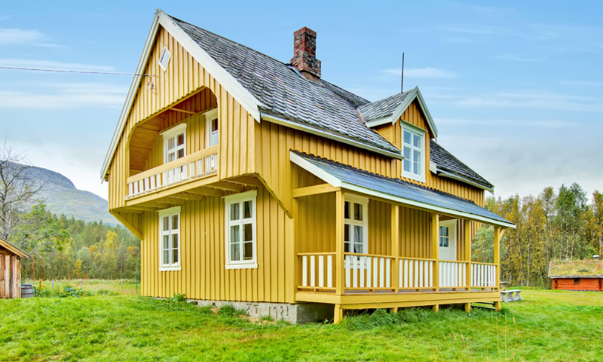 Slette med gress og et stort, gult, gammelt hus. Åser og fjell i bakgrunnen, samt blå himmel. None mindre bygninger i bakgrunnen til venstre og høyre.