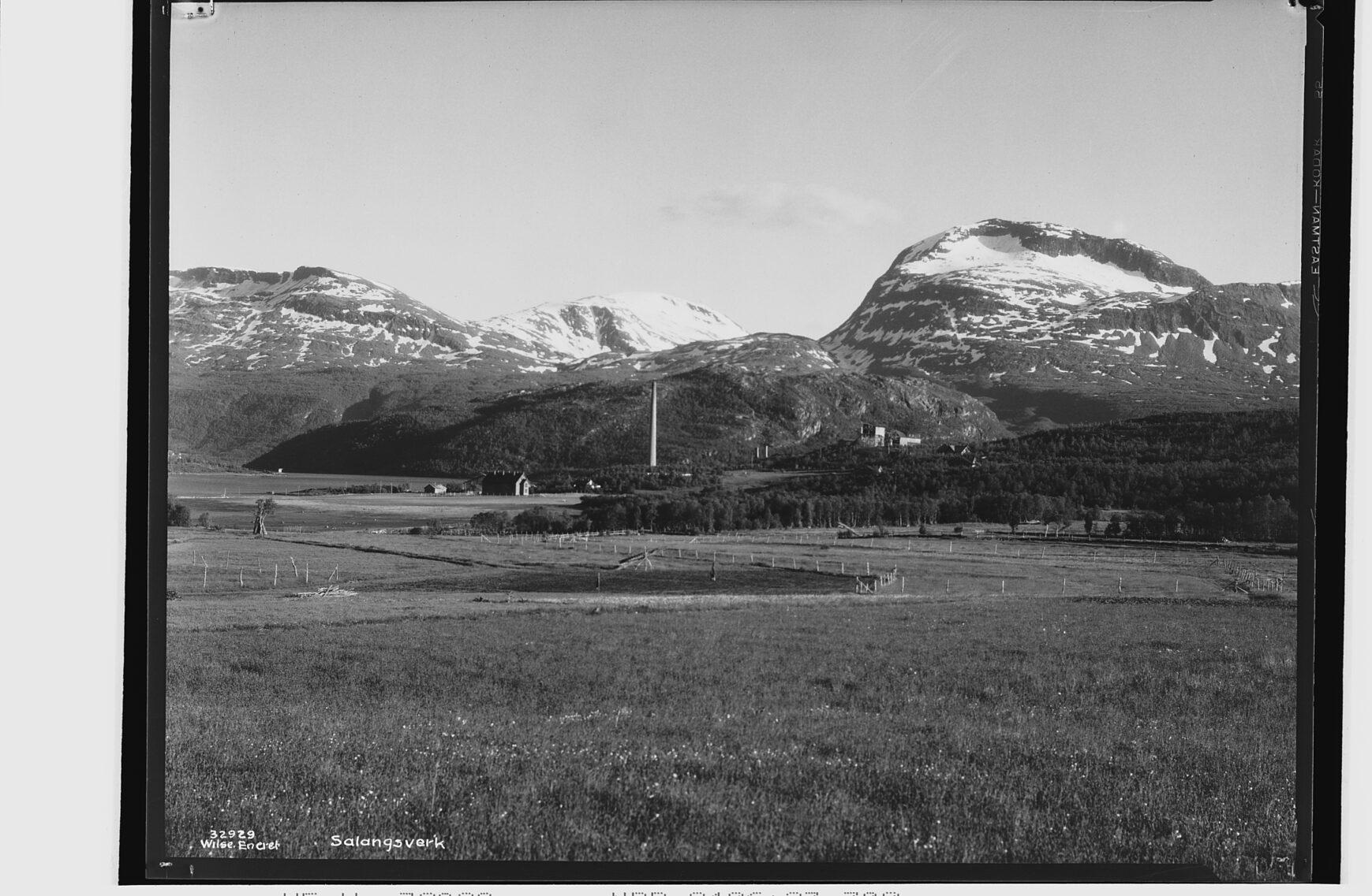 Svart hvitt bilde. Et stort gjorde i forgrunnen og pipa og noe av verket midt i. Snøkledde fjell i bakgrunnen.