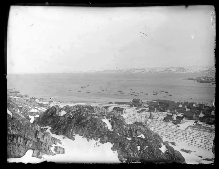 Et eldre svart‑hvitt oversiktsfoto tatt fra en høyde. I forgrunnen ligger steinete terreng med flekker av snø, og lenger nede ses en kystbebyggelse med små trebygninger og tørkestativer. Ute på sjøen ligger en rekke små båter spredt over vannflaten. I bakgrunnen skimtes fjell og landformer på den andre siden av fjorden.