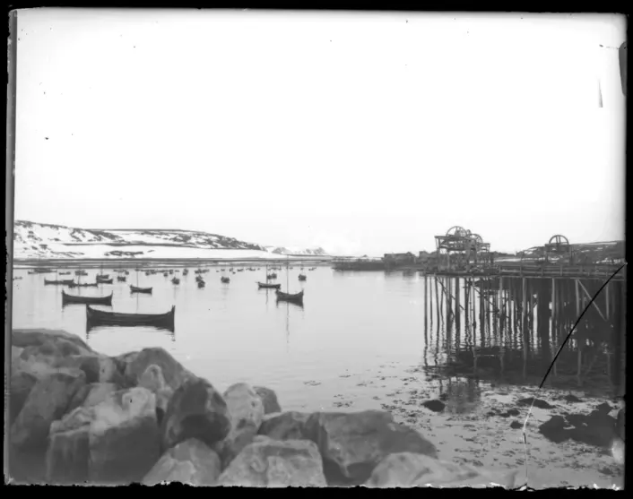 Et historisk svart‑hvitt foto av en rolig bukt med små trebåter som ligger fortøyd i vannet. I forgrunnen ligger store steiner ved strandkanten, og til høyre står en enkel bryggekonstruksjon på påler. I bakgrunnen ses snødekte åser under en lys, overskyet himmel.