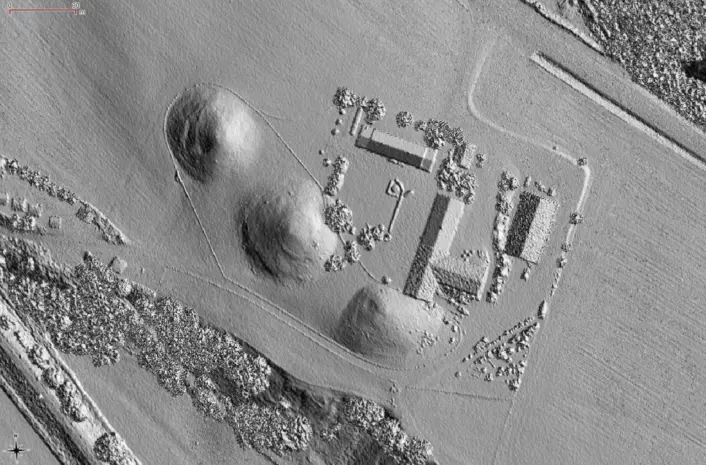 «Et gråtonebilde som viser et landskap sett ovenfra, med tre markante hauger ved siden av et rektangulært bygningskompleks. Terrenget rundt består av åpne flater og områder med trær langs kantene.»