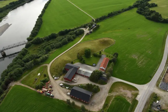 «Luftfoto av et gårdsanlegg omgitt av store grønne jorder. En elv renner langs venstre side av bildet, med en liten bro og en gårdsvei som slynger seg inn til bygningene. Flere røde og mørke bygninger står samlet rundt et gårdstun, og veier og trær omkranser området.»