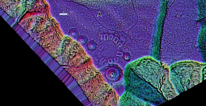 «Et fargerikt topografisk visualiseringskart som viser et område med ujevnt terreng. I midten ligger en gruppe sirkulære forhøyninger og groper arrangert i et mønster. Området rundt vises i kontrastfarger i lilla, blått, grønt og rødt, som fremhever høydeforskjeller. Kartet er vist i skrå vinkel med en markert svart bakgrunn langs to kanter.»