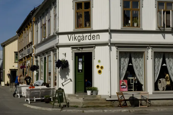 Et gatehjørne med en hvitmalt trebygning med skiltet «Vikgården». Bygget har store vinduer med blondegardiner, en grønn inngangsdør og blomsterdekorasjoner. Utenfor står utemøbler, pynt og ulike varer på display, og flere bygninger i tre ligger langs gaten i bakgrunnen.