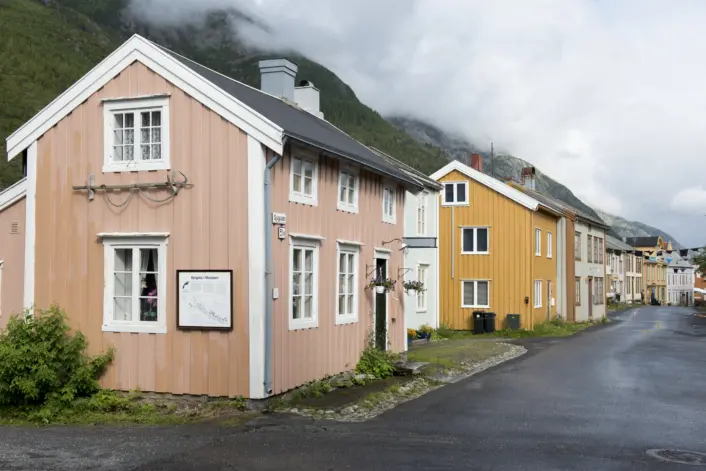 Et lite trehus malt i lys rosa med hvite karmer står nær en asfaltert vei, omgitt av flere trebygninger i gule og hvite farger. Husene ligger tett sammen i et fjordlandskap med grønne fjellsider og lave skyer i bakgrunnen. Veien foran bygningene er våt, og vegetasjon vokser langs kanten av det rosa huset.