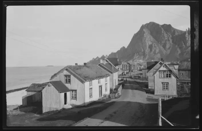 Svart/hvitt bilde. Grusvei med små trehus på hver side. Bratt fjell i bakgrunnen. Havet ute til venstre.