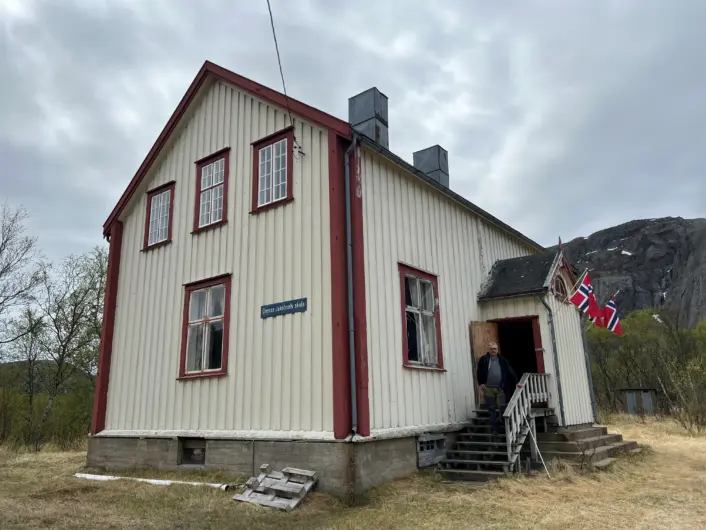 "Et hvitt trehus med røde detaljer og en trapp opp til inngangen. Bygningen har et skilt med teksten 'Grense Jakobselv' og norske flagg henger på utsiden. I bakgrunnen sees fjell og trær under en overskyet himmel. En mann står på trappa"