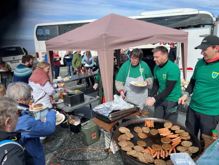 «Flere mennesker står rundt en utendørs matbod der det lages mat. På et stort grillfat ligger mange burgere til steking, mens personer bak boden serverer mat. I bakgrunnen står en buss og flere mennesker, og over boden er det satt opp et lite telt under en klar blå himmel.»