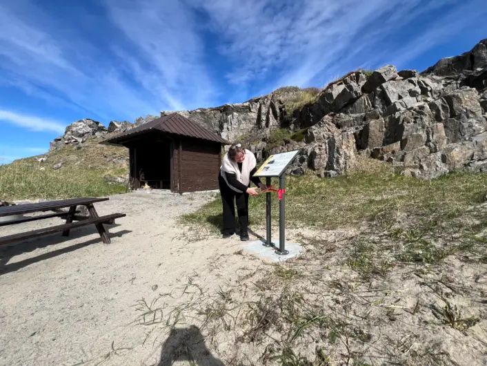 «En person står ved et informasjonsskilt i et åpent område med gress og stein. Like ved står en liten mørk bygning og en benk. I bakgrunnen er det bratte, steinete fjell under en klar blå himmel med lette skyer.»