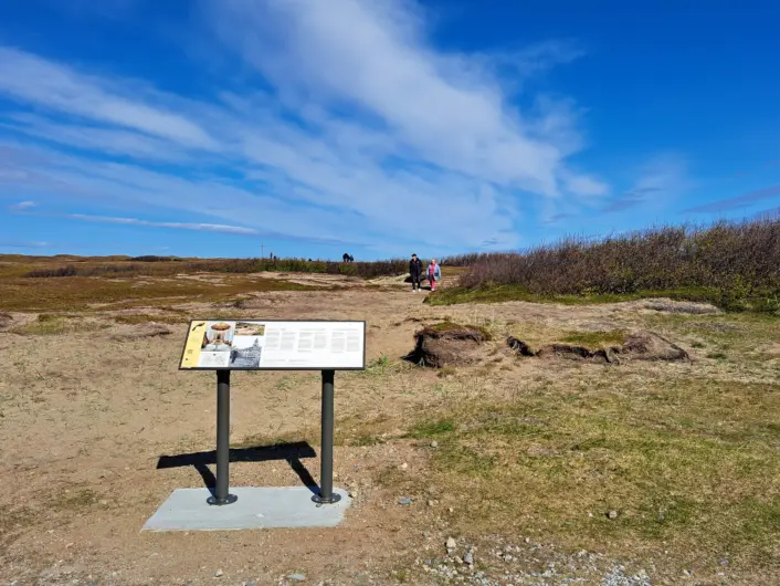 «Informasjonsskilt står på en grusplass i et åpent landskap med gress og lave busker. En sti går videre innover i terrenget, og himmelen er klar og blå med lette skyer.»