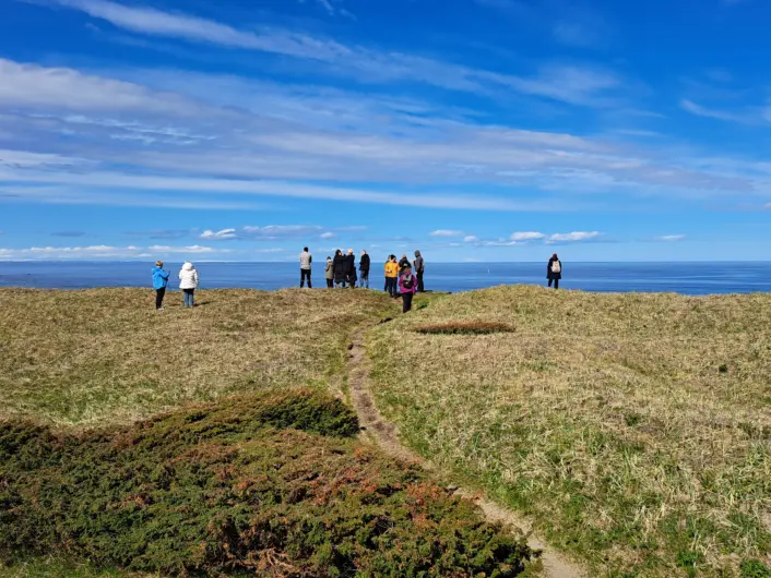 «En smal sti går gjennom et åpent gresslandskap mot en gruppe mennesker som står på en liten høyde. I bakgrunnen ses havet under en klar blå himmel med spredte skyer.»