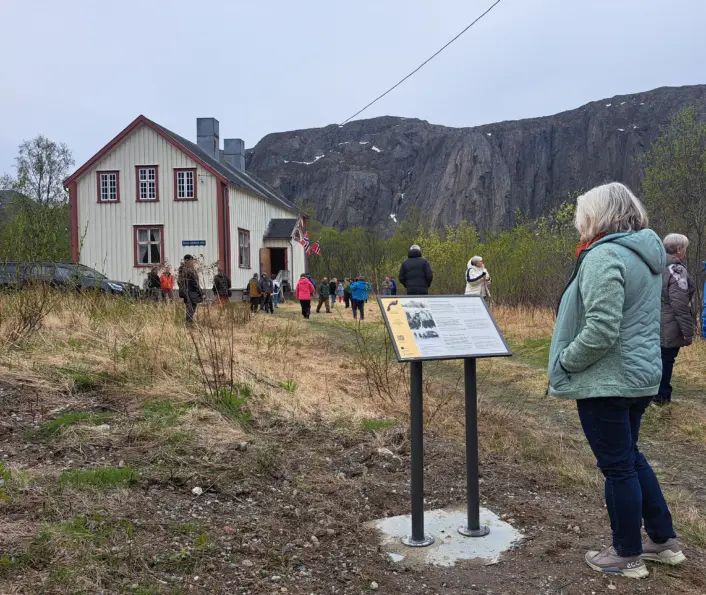 Hvit trebygning med røde karmer i bakgrunnen. Overskyet vær. Flere mennesker foran huset og helt fremst i bildet et informasjonsskilt og en kvinne som leser på det.