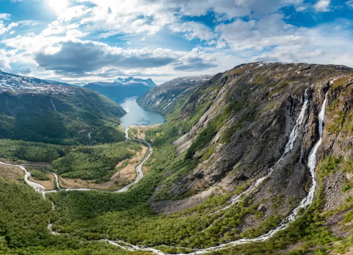 Panoramautsikt over et fjordlandskap omgitt av bratte fjellsider. En svingete vei slynger seg gjennom den grønne dalen nedenfor, mens en høy foss renner ned langs fjellveggen til høyre. I bakgrunnen strekker den blå fjorden seg mellom fjellene under en himmel med spredte skyer.