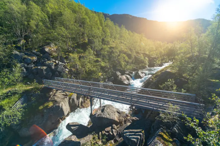 Solbelyst landskap med en smal bro som krysser en fossende elv mellom store steiner. Broen er omgitt av grønn skog og fjellterreng, mens sollyset skinner inn fra øvre høyre del av bildet og gir et varmt preg på naturen.