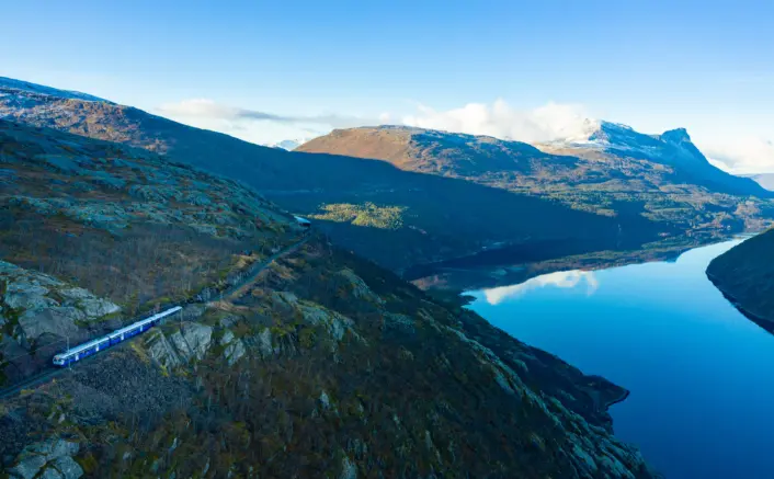 Tog på jernbanespor langs en fjellside ved en stille innsjø. Landskapet består av nakne fjell, lav vegetasjon og klart vann som speiler fjellene og den blå himmelen. Toget følger dalsiden med utsikt over innsjøen og fjellene i bakgrunnen.
