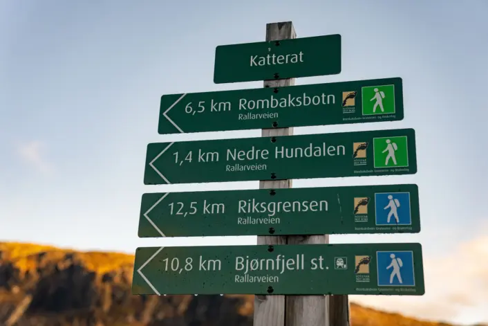 Grønt turveiskilt i fjellandskap med avstander til Katterat, Rombaksbotn (6,5 km), Nedre Hundalen (1,4 km), Riksgrensen (12,5 km) og Bjørnfjell stasjon (10,8 km).