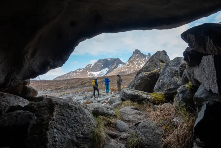 Utsikt fra fjellhule mot åpent fjellandskap. Tre personer står rett utenfor hula. Spisse fjell og blå himmel i bakgrunnen.