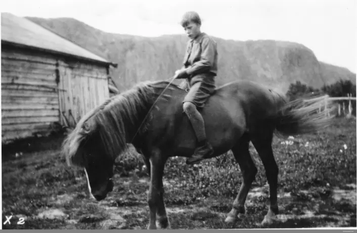 Svart hvitt bilde. Gutt sitter på hest. Fjøs eller lignende bygning til venstre i bakgrunnen. Fjell helt bak.