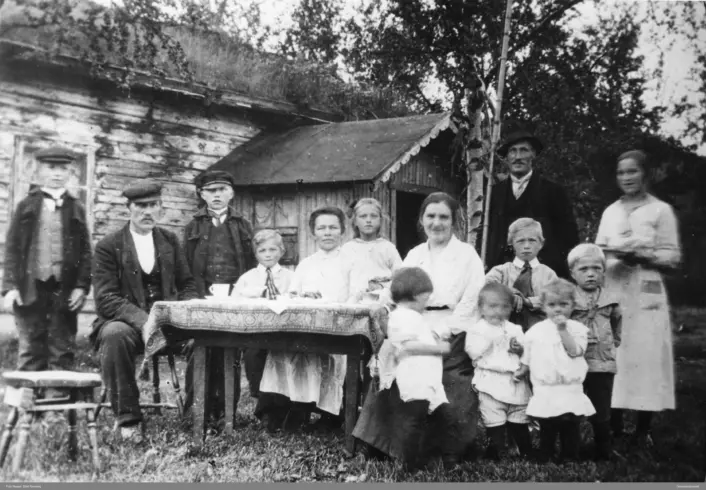 Svart hvitt bilde. Familie bestående av voksne og barn, totalt 14 mennesker sitter og står foran et gammelt hus med torvtak. Et bord foran de som sitter.