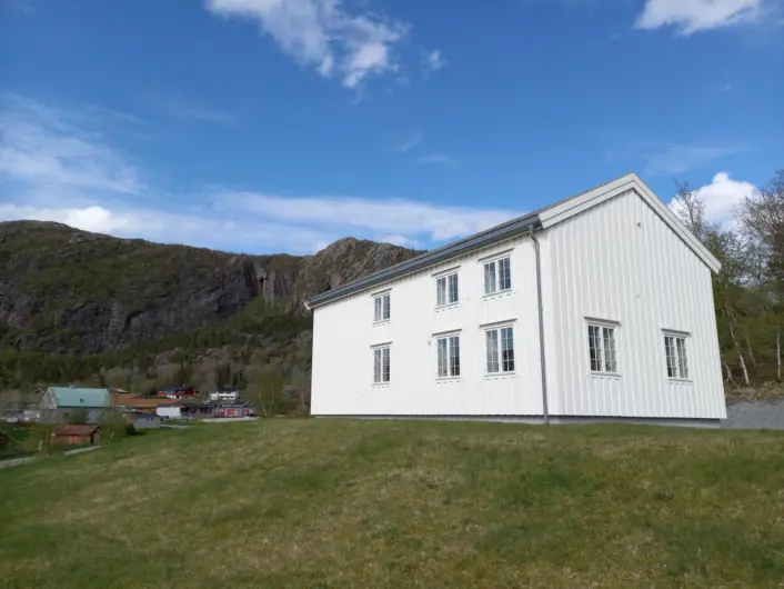 Hvitt hus på en liten gresskledt haug. Ås i bakgrunnen, blå himmel