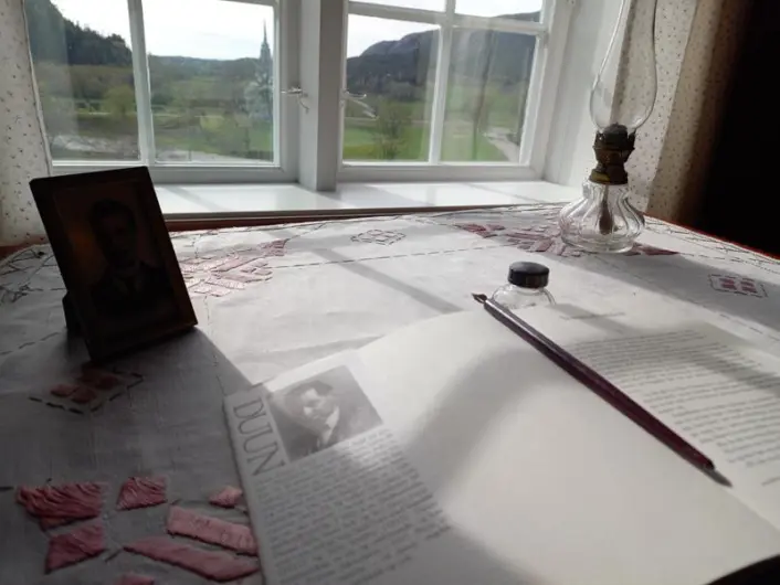 Åpen skrivebok med blyant på en pult som står ved vinduet. Utsikt mot åpent landskap utenfor vinduet.