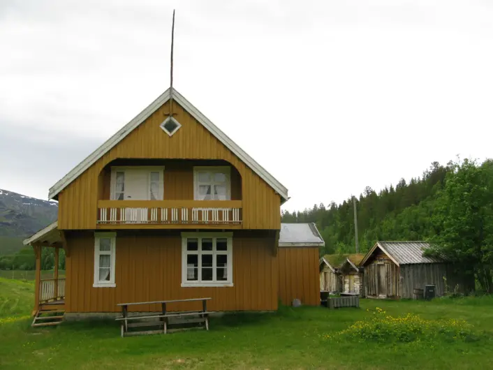 En gressplen og et stort, gult gammelt hus i midten. Noen mindre hus kimtes bak til høyre. Åser til høyre og venstre i bakgrunnen og en himmel med tett skydekke.
