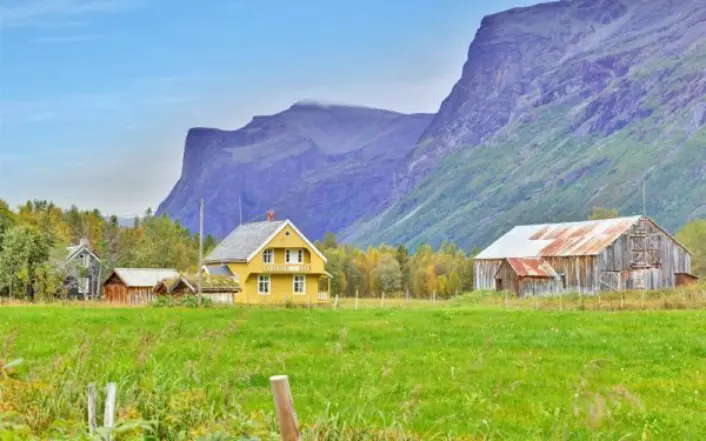 En grønn eng i forgrunnen og så et gult, gammelt hus og en nedslitt fjøs til høyre. En mindre bygning bak det gule huset. Noen trær bak husene og et høyt fjell i bakgrunnen. Blå himmel.