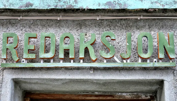 Slitt, grønn metallskrift som danner ordet ‘REDAKSJON’ på en værbitt fasade over en dør. Bokstavene er delvis rustne og står på en gammel, grå betongvegg.