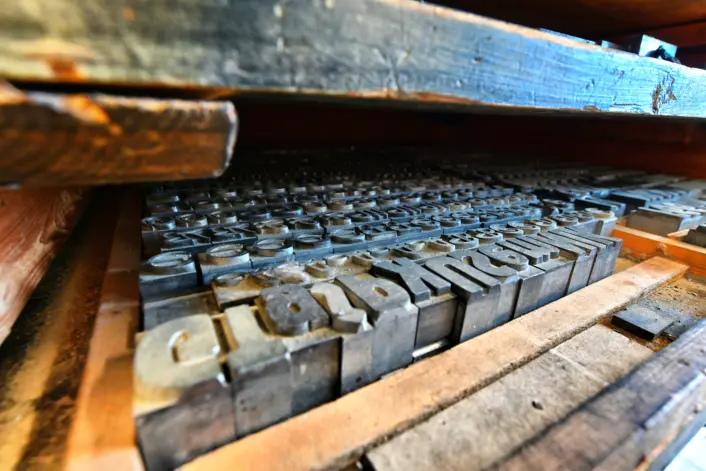 Nærbilde av en skuff med gamle blytyper til trykking. De små metallbokstavene ligger tett i tett i et slitt trekabinett, delvis skjult av en hylle over.