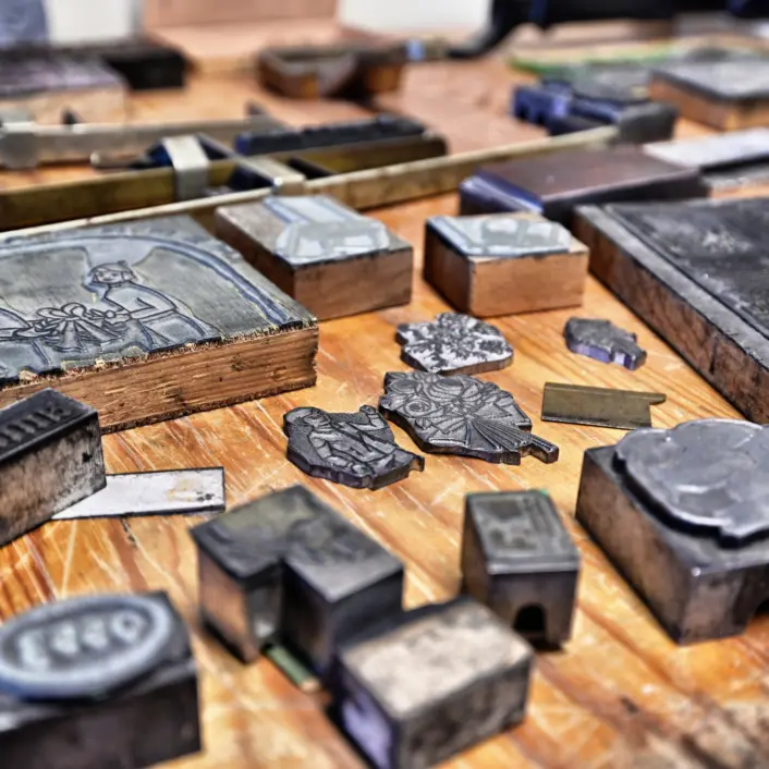 Ulike metallklisjeer og trykkblokker ligger spredt utover en trebordflate. Gjenstandene har inngravert tekst og motiver, og viser bruksspor fra tradisjonelt trykkehåndverk.