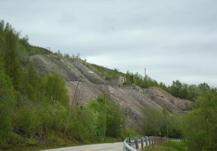 En vei går gjennom et grønt dalområde med trær på begge sider. På skråningen ovenfor veien ligger flere store stein- og grusflater som ser ut som gamle tipphauger eller terrenginngrep, delvis omkranset av vegetasjon. Himmelen er overskyet.