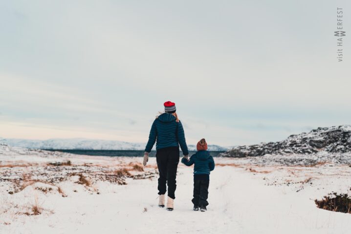 Vinterlandskap med snø. En voksen dame og et barn går hånd i hånd med ryggen til ut mot havet. De går på en slette og foran dem kan en se et snødekt berg til høyre og havet til venstre.