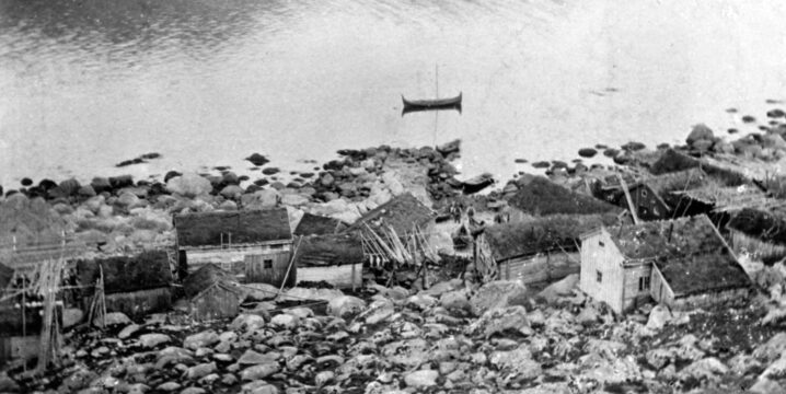 Svart hvitt bilde. Fiskevær sett ovenfra. Fjorden foran med en Nordlandsbåt liggende på vannet.