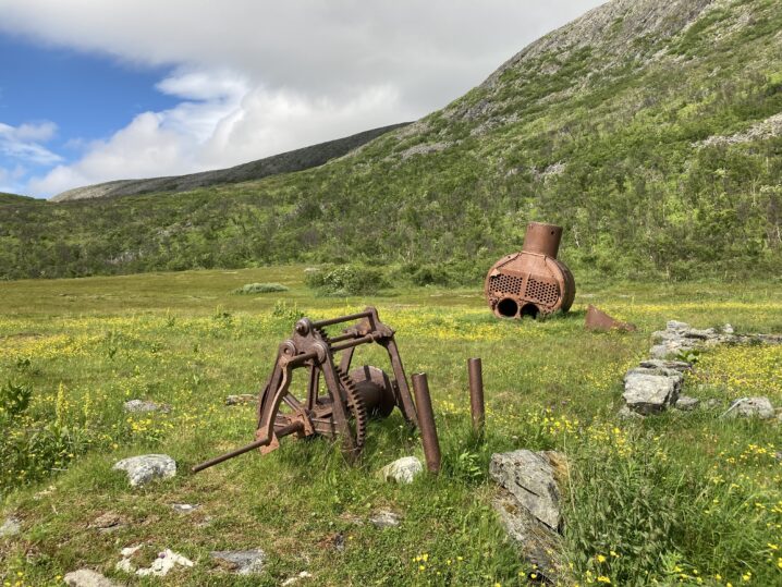 Grønn eng med et lavt fjell i bakgrunnen. På enga ser en en rustet dampkjele og spill.