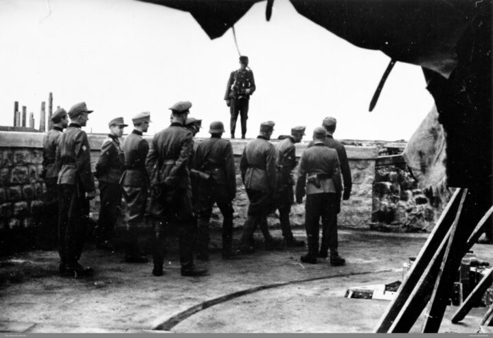 11 menn i tysk uniform med ryggen til. En soldat i uniform står vendt mot de andre oppå en mur. Hvit himmel over.