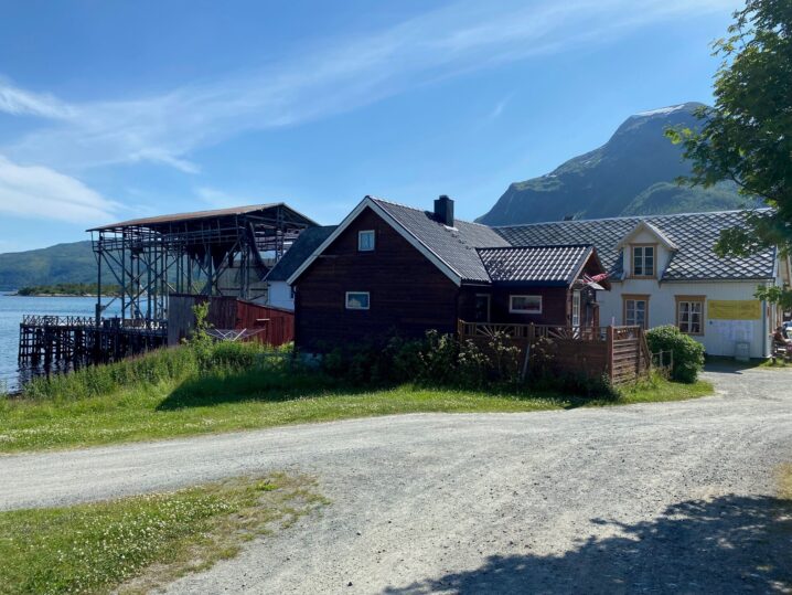 Grusvei i forgrunnen. Et rødt og et hvitt gammelt hus ved en brygge. Fjell og fjord i bakgrunnen.