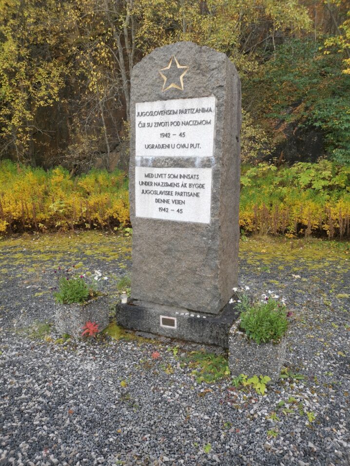 "Minnesmerke i stein med inskripsjonsplate, plassert på en gruslagt plass. Rundt monumentet står planter og høstfargede trær i bakgrunnen."