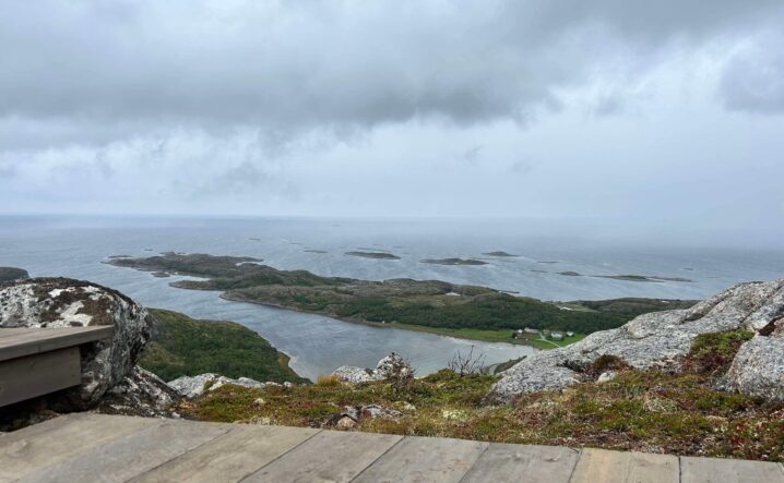 Utsikt fra et høyt punkt mot et kystlandskap med mange lave, grønne øyer og holmer omgitt av hav. I forgrunnen ligger en treplatting og steiner, mens horisonten og himmelen er dekket av grå skyer som skaper et overskyet og noe disig inntrykk.