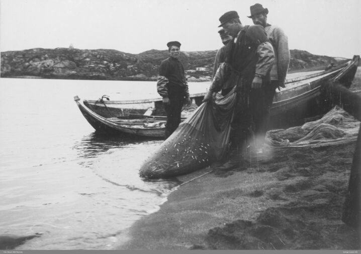 Fiskere som drar garn . Står på en strand. Robåt i bakgrunnen. Berg helt bakerst.