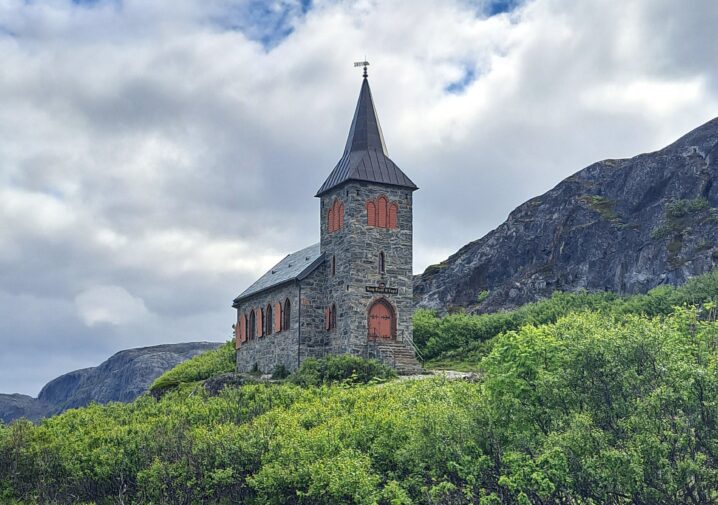 Kapell bygget i stein står på en høyde med mye grønn vegetasjon. Fjell i bakgrunnen. Himmel med hvite skyer.