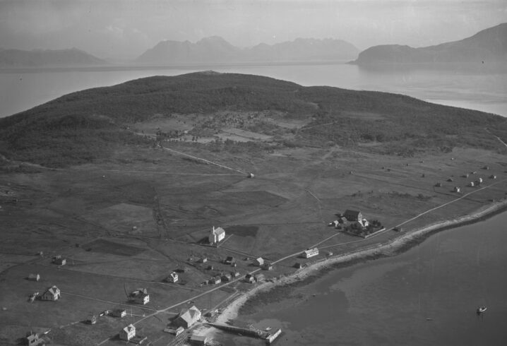 Svart hvitt bilde. Flyfoto. Spredt bebyggelse på en øy. Hav og fjell i bakgrunnen.