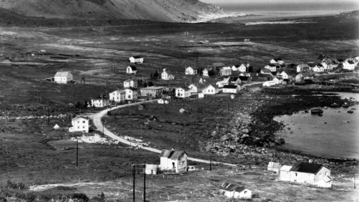 Svarthvitt-bilde av et lite fiskevær med en rekke hvite hus langs en vei. Åpent landskap bak til venstre og fjære og noe hav til høyre.
