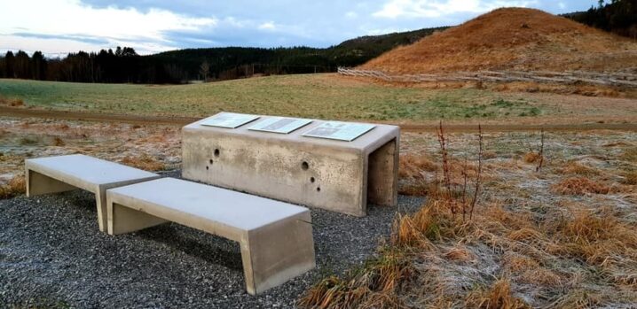 På et flatt område med grus og gress står to benker i betong foran en lav installasjon med skilt. Foran ser en den ene av gravhaugene på Bertnem. Bak der noen åser og blå himmel med skyer.