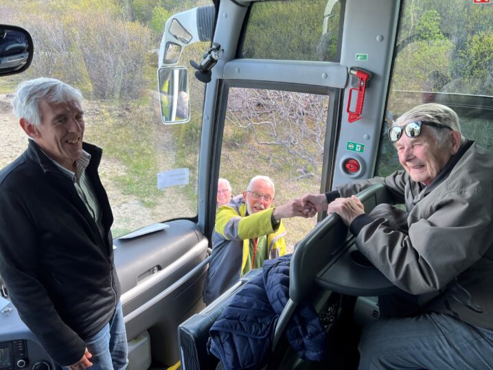 "En eldre mann sitter i en buss og snakker med en annen person, mens en tredje person i bakgrunnen ser ut av vinduet. Utsikten viser et landskap med trær og fjell."