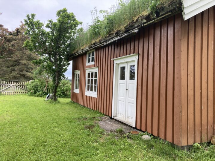 Dette bildet viser en tradisjonell trebygning med brune, vertikale panel. Bygningen har en hvit dør og et vindu med hvite rammer. Taket er dekket av gress og vegetasjon, noe som indikerer at det kan være et grønt tak designet for å integrere seg med naturen. Rundt bygningen er det en velholdt plen med grønt gress, og i bakgrunnen kan man se trær og et fjellandskap under en skyet himmel.