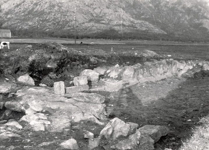 Svart hvitt bilde . Ruiner av kirke i forgrunnen. Åpent område bak og så nederste del av av grått fjell i bakgrunnen.