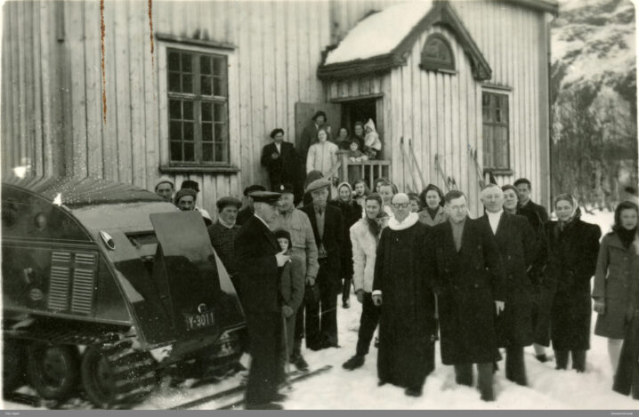 Svart hvitt bilde. Gamme skolebygning malt i hvitt i bakgrunnen. Folkemengde foran, samt en bil til venstre. Snø på bakken.