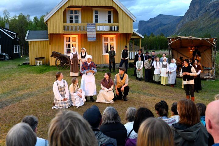 "En utendørsspel foran et gult trehus i norsk landskap. En gruppe mennesker i historiske klær spiller teater, mens et publikum sitter på gresset og ser på."