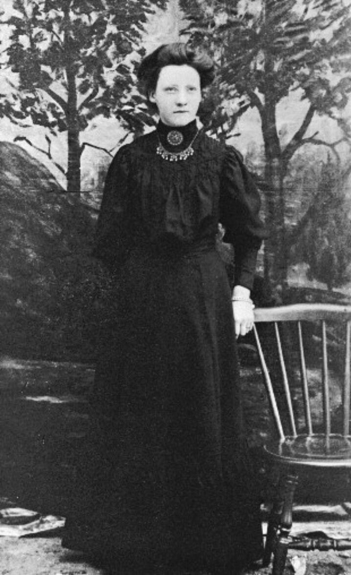 Svart-hvitt fotografi av en stående person i en lang, mørk kjole. Personen står ved siden av en stol i et atelier med malt bakgrunn som forestiller trær. Fotografiet er et eldre portrett, trolig fra slutten av 1800-tallet eller begynnelsen av 1900-tallet.